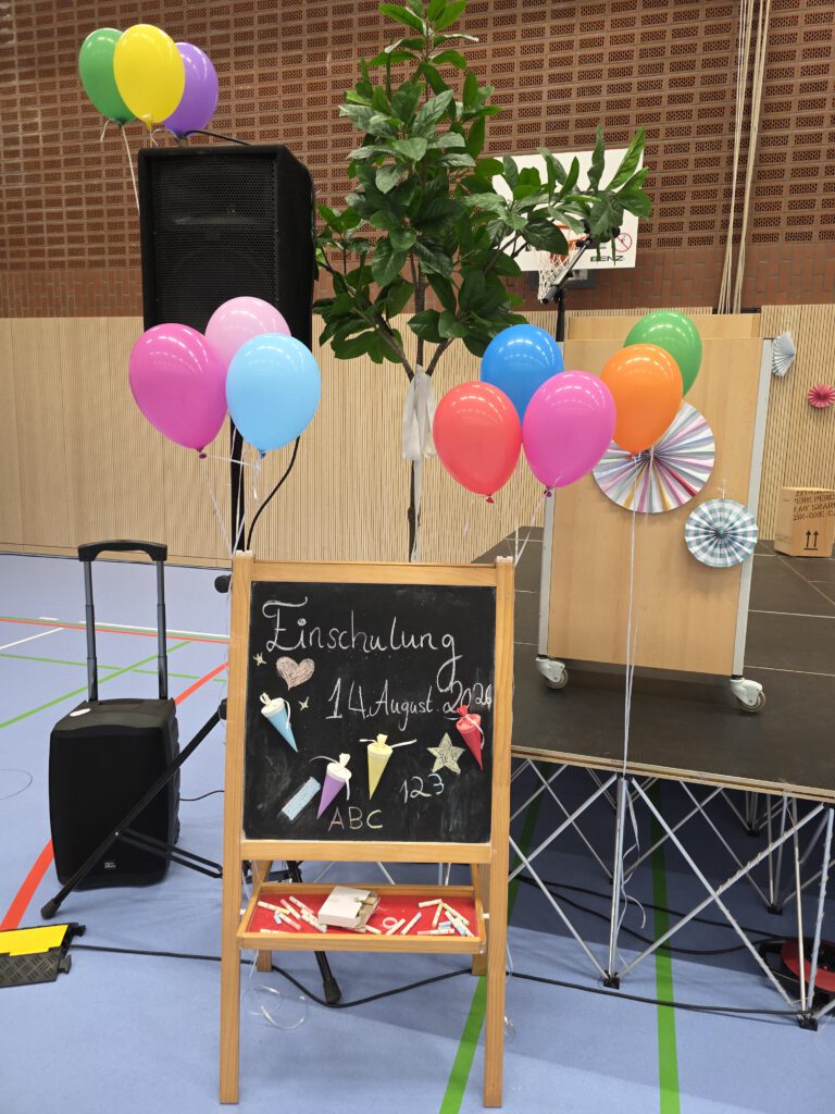 Kleine Holztafel mit der Aufschrift "Einschulung14. August 2026". Auf der Tafel befinden sich kleine aufgeklebte Schultüten. Links von der Tafel befindet sich eine Musikbox, im Hintergrund ist eine aufgebaute Bühne mit einem Rednerpult und Luftballons zu sehen.