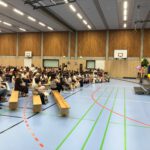 Menschenmenge sitzt in einer Turnhalle auf Holzbänken und hört einer Rede des Schuleiters zu, der rechts im Bild auf einer Bühne steht