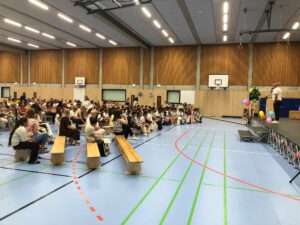 Menschenmenge sitzt in einer Turnhalle auf Holzbänken und hört einer Rede des Schuleiters zu, der rechts im Bild auf einer Bühne steht