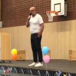 Der Schulleiter steht mit einem Mikrofon auf der Bühne und hält eine Willkommensrede auf der Einschulungsfeier für den 5. Jahrgang.