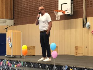 Der Schulleiter steht mit einem Mikrofon auf der Bühne und hält eine Willkommensrede auf der Einschulungsfeier für den 5. Jahrgang.