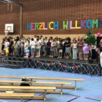 Kinder und Lehrkräfte stehen auf der Bühne einer Turnhalle. Im vorderen Bereich des Bildes sind Holzbänke zu sehen. Auf der rechten Seite steht der Schulleiter mit einem Mikrofon in der Hand.