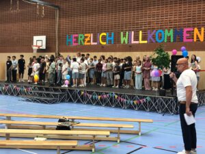Kinder und Lehrkräfte stehen auf der Bühne einer Turnhalle. Im vorderen Bereich des Bildes sind Holzbänke zu sehen. Auf der rechten Seite steht der Schulleiter mit einem Mikrofon in der Hand.